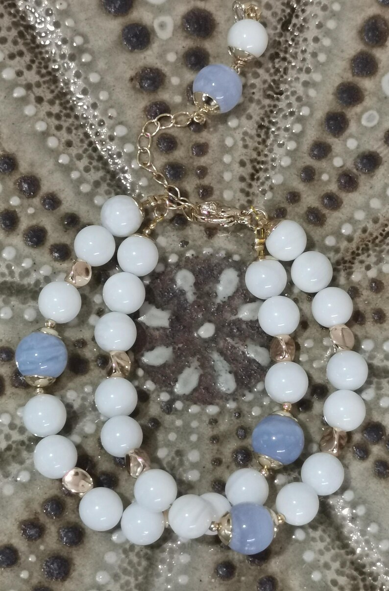 Pu&ograve; includere: Un set di collana e bracciale con perline bianche e azzurre. La collana ha una sottile catena d'argento, e entrambi i pezzi hanno dettagli color oro. I gioielli sono esposti su una superficie strutturata dai toni neutri.