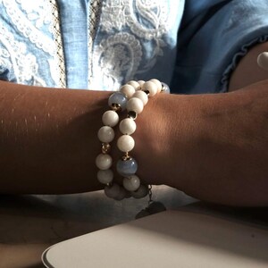 Pu&ograve; includere: Due braccialetti di perline con perline bianche e azzurre. I braccialetti sono indossati su un polso, con una camicia azzurra sullo sfondo. I braccialetti hanno dettagli dorati e un piccolo ciondolo.