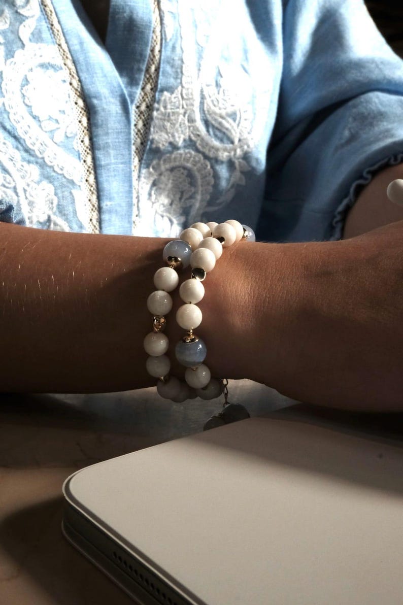 Pu&ograve; includere: Due braccialetti di perline con perline bianche e azzurre. I braccialetti sono indossati su un polso, con una camicia azzurra sullo sfondo. I braccialetti hanno dettagli dorati e un piccolo ciondolo.