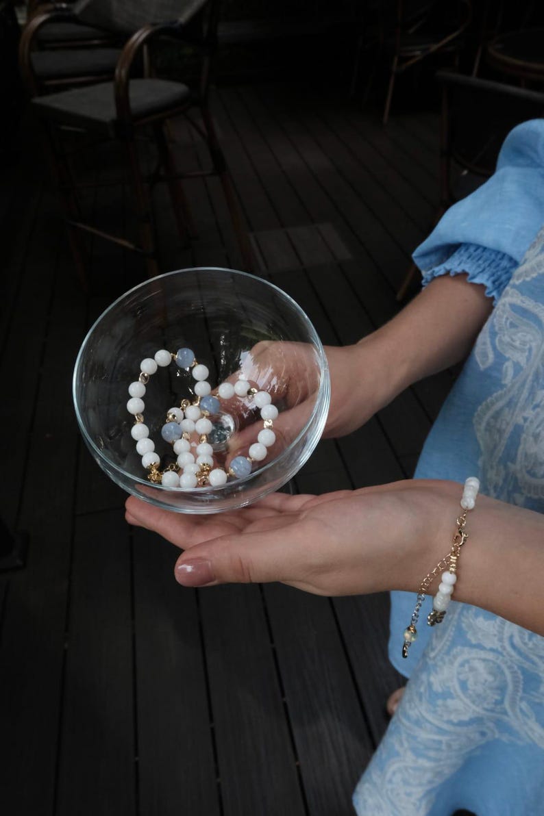Pu&ograve; includere: Una ciotola di vetro trasparente contiene una collana di perline bianche con dettagli dorati e perline azzurre. Un braccialetto abbinato &egrave; indossato al polso. I gioielli sono esposti su uno sfondo di legno scuro.