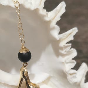 May include: A gold-coloured high-heeled shoe charm hangs from a delicate gold chain, with a black bead and gold cap accent. The charm is set against a white, textured background.