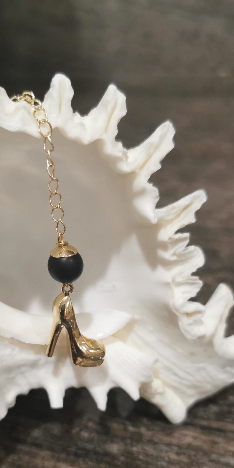 May include: A gold-coloured high-heeled shoe charm hangs from a delicate gold chain, with a black bead and gold cap accent. The charm is set against a white, textured background.