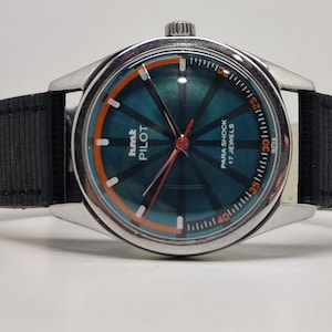 Puede incluir: Reloj HMT Pilot vintage con esfera color turquesa y detalles en naranja. La caja es plateada y la correa es de tela negra. La esfera muestra el texto "PARA SHOCK 17 JEWELS INDIA".
