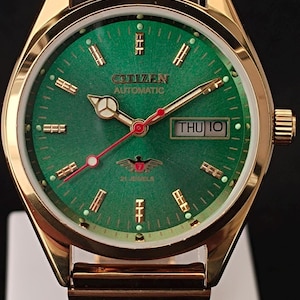 May include: Gold-toned Citizen automatic watch with a green dial. The watch features gold hands and markers, a day-date display, and a gold link bracelet. The watch face displays the text "Citizen Automatic" and "21 Jewels".