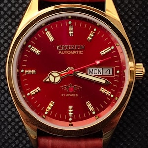 Pode incluir: Um relógio de pulso automático Citizen dourado com mostrador vermelho profundo e pulseira de couro a condizer. O mostrador do relógio exibe o dia e a data, com o dia a indicar "MON 4". O relógio tem ponteiros e marcadores dourados, e a pulseira é vermelha rica e texturizada.