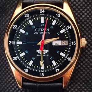 Puede incluir: Un reloj Citizen Automatic dorado con esfera negra y correa de cuero negra. El reloj presenta números blancos, un segundero rojo y una pantalla de fecha. La esfera del reloj dice "CITIZEN AUTOMATIC".
