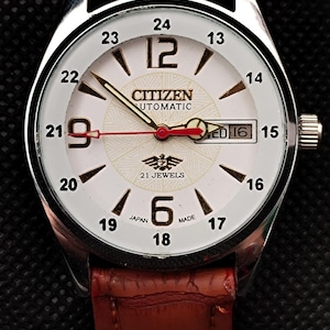 May include: A Citizen automatic wristwatch with a white face, black numerals, and a brown leather strap. The watch displays the day and date, with the text "21 JEWELS" and "JAPAN MADE".