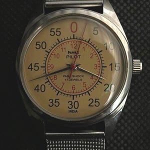 May include: A vintage wristwatch with a silver case and mesh strap. The watch face is tan with black and red numerals, and the text "PILOT" and "INDIA" are visible. The watch has a second hand.