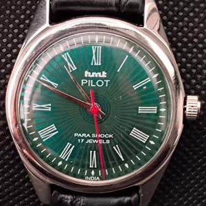 May include: A silver wristwatch with a green dial, white Roman numerals, and red hands. The watch face shows "hmt PILOT" and "PARA SHOCK 17 JEWELS". It has a black leather strap. The watch is made in India.
