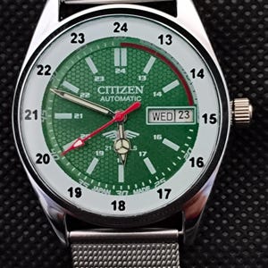 Pode incluir: Relógio de pulso automático Citizen com mostrador verde e pulseira de malha prateada. O mostrador mostra o dia e a data, com ponteiros e marcadores vermelhos. Fabricado no Japão.