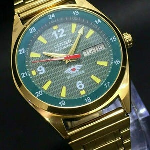 Op de afbeelding: Goudkleurig Citizen automatisch horloge met een groene wijzerplaat en gouden wijzers. De wijzerplaat toont de dag en datum, met gele uurmarkeringen en een gouden lunette. De horlogeband is ook goudkleurig.