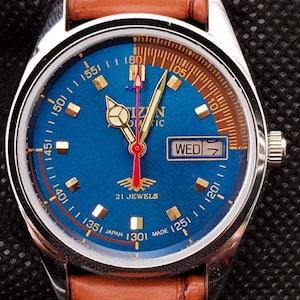 Könnte beinhalten: Eine Vintage-Armbanduhr mit blauem Zifferblatt, goldenen Zeigern und einem braunen Lederarmband. Das Zifferblatt zeigt den Wochentag und den Text "21 JEWELS". Die Uhr ist mit "JAPAN MADE" gekennzeichnet.