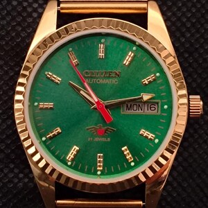 Può includere: Orologio automatico Citizen color oro con quadrante verde. L'orologio presenta un cinturino a maglia e un display della data che mostra "LUN 16". Il quadrante include il testo "CITIZEN AUTOMATIC" e "21 JEWELS".