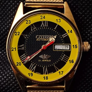 May include: Gold-toned Citizen automatic wristwatch with a yellow and black face. The watch features Roman numerals, a date display, and a mesh band. The watch face displays the text "Citizen Automatic 21 Jewels".