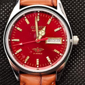 Peut inclure: Montre-bracelet automatique Citizen avec cadran rouge et aiguilles et index dorés. La montre affiche le jour et la date et est montée sur un bracelet en cuir marron. Le cadran indique "Citizen Automatic" et "21 Jewels".
