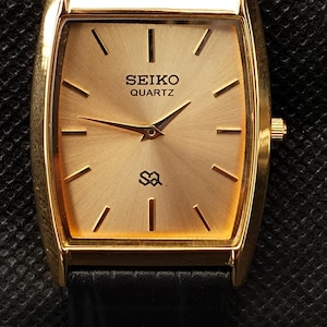 Puede incluir: Un reloj Seiko Quartz dorado con una esfera rectangular y una correa de cuero negro. La esfera del reloj muestra el logotipo de Seiko y la palabra "QUARTZ". El reloj tiene manecillas e indicadores de hora dorados.