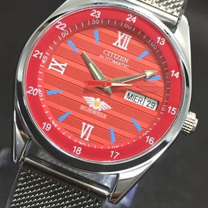 Könnte beinhalten: Eine Citizen Automatic Uhr mit rotem Zifferblatt und silbernem Mesh-Armband. Das Zifferblatt hat römische Ziffern, blaue Markierungen und eine Datumsanzeige. Das Gehäuse ist aus Edelstahl.