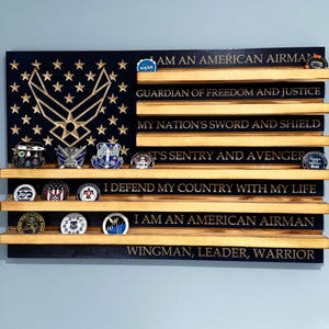 May include: A wooden American flag-shaped display with shelves for challenge coins. The flag features the Air Force symbol and stars, with phrases like "I AM AN AMERICAN AIRMAN" and "GUARDIAN OF FREEDOM AND JUSTICE". Various challenge coins are displayed.