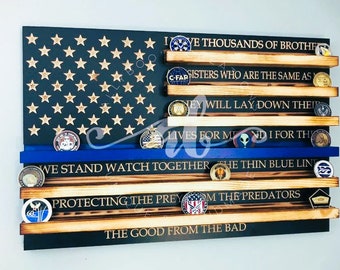 May include: A wooden American flag with a blue stripe and five shelves for displaying challenge coins. The flag has gold stars on a dark blue background. The shelves have text on them, including "We Stand Watch Together The Thin Blue Line", "Protecting The Prey From The Predators", and "The Good From The Bad".