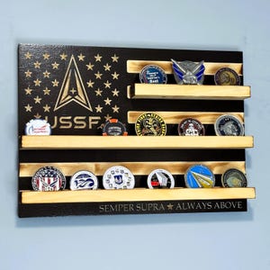 May include: Wooden American flag-shaped display shelf with three rows for challenge coins. The flag design features stars, stripes, and the USSF logo. The bottom reads "Semper Supra Always Above."
