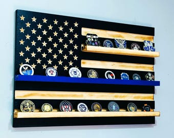 May include: A wooden American flag with a blue stripe, featuring shelves for displaying challenge coins. The flag is painted black with gold stars and has a natural wood finish on the stripes.