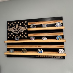 May include: Wooden American flag-shaped display shelf with multiple shelves holding various challenge coins. The flag design features a black field with stars and a military emblem, and the text "PRIDE RUNS DEEP".