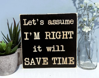 May include: A square black wooden sign with the text "Let's assume I'M RIGHT it will SAVE TIME" in gold lettering. The sign is displayed on a white shelf with a small succulent plant and a glass vase with white flowers.