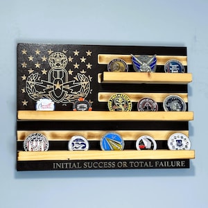 May include: Wooden challenge coin display in the shape of an American flag. The display has black stripes and wooden shelves holding various coins. The top section features a gold emblem and stars. The bottom reads "INITIAL SUCCESS OR TOTAL FAILURE."
