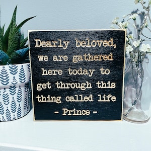 May include: Black wooden sign with gold lettering that reads "Dearly beloved, we are gathered here today to get through this thing called life - Prince -"