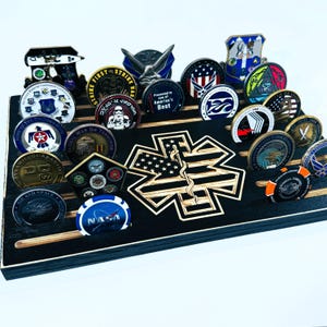 May include: A black wooden display stand with a star of life design and a row of 20 military challenge coins. The coins feature various military branches, including the Air Force, Army, and Navy. The stand is designed to hold and display challenge coins.