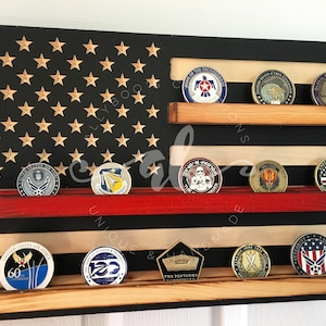 May include: A wooden American flag display with three shelves for displaying challenge coins. The flag is painted black with gold stars and red and white stripes. The shelves are stained in a natural wood color.