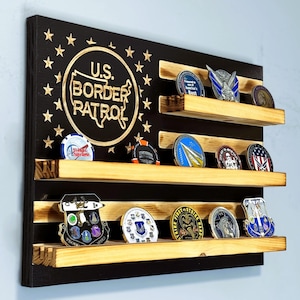 May include: A black wooden coin display with three shelves, holding various challenge coins. The display features the text "U.S. Border Patrol" in a gold circle, surrounded by gold stars. The shelves are made of natural wood.