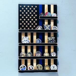May include: Wooden American flag display with shelves, featuring a blue stripe and star cutouts. The shelves hold various challenge coins and pins. The flag design is made of black and natural wood, creating a patriotic theme.