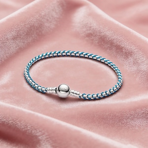 May include: A silver bracelet with a woven design and a blue accent. The bracelet has a round clasp with the brand name "Pandora" engraved on it. The bracelet is displayed on a pink satin surface.