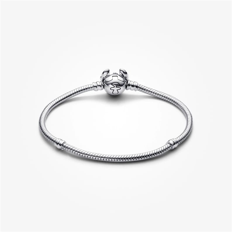 May include: A silver bracelet with a round clasp featuring a decorative design. The bracelet has a snake chain design and is displayed on a white background. The bracelet is a piece of jewellery.