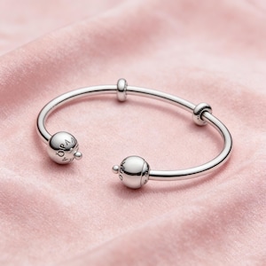 Könnte beinhalten: Ein silberfarbenes Armband mit zwei kugelförmigen Endkappen und zwei kleineren dekorativen Perlen. Das Armband wird auf einem rosa, seidigen Stoff präsentiert. Die Endkappen tragen die Gravur "OPA".