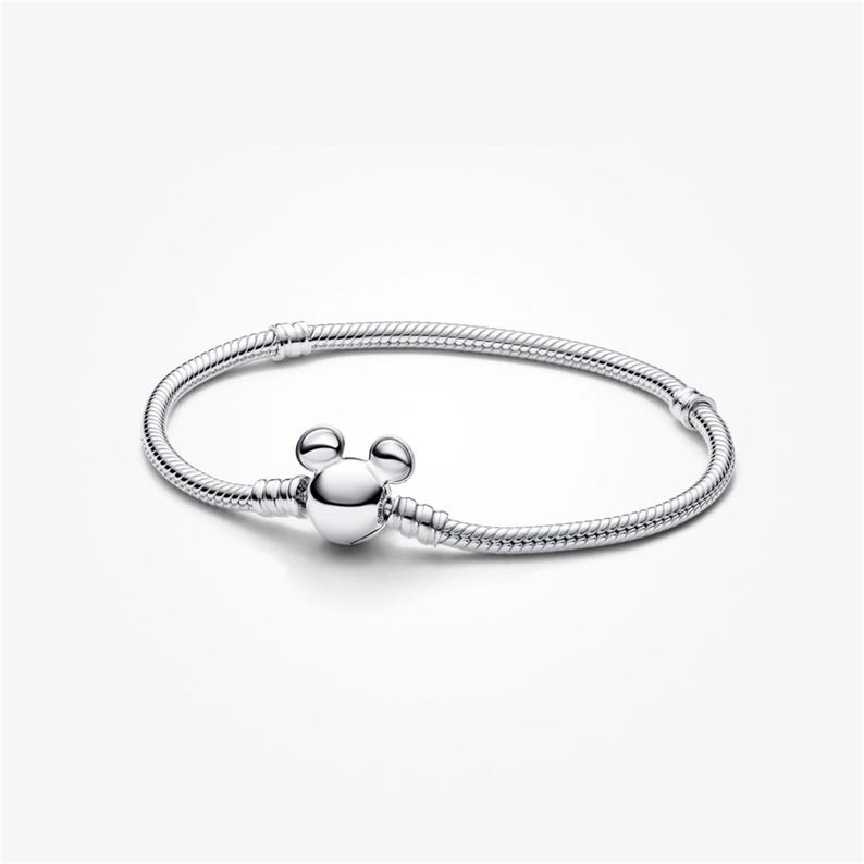 May include: A silver bracelet with a Mickey Mouse-shaped clasp. The bracelet is made of a flexible, snake-like chain. The clasp is a round, polished silver charm with two small round accents above it, resembling Mickey Mouse ears.