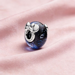 May include: A silver charm with a dark blue and black design, featuring a bow and two small circular accents. The charm has a spherical shape and is likely part of a jewelry collection. The charm has the word "Disney" on it.