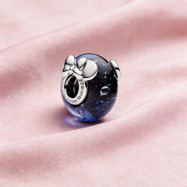 May include: A silver charm with a dark blue and black design, featuring a bow and two small circular accents. The charm has a spherical shape and is likely part of a jewelry collection. The charm has the word "Disney" on it.