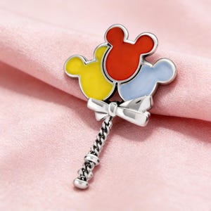 K&ouml;nnte beinhalten: Eine silberfarbene Brosche in Form eines Mickey Mouse-Ballon-Bouquets. Die Ballons sind rot, gelb und hellblau. Die Brosche hat eine Schleife und einen kettenartigen Griff. Der Hintergrund ist ein rosa Stoff.