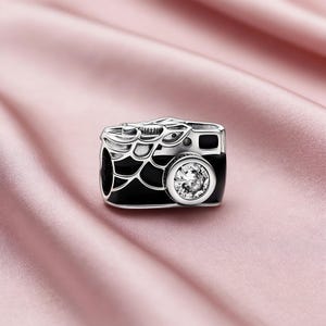 May include: A silver-toned charm in the shape of a vintage camera. The camera body is black with silver detailing and a large, clear, round gem at the lens. The charm is set against a soft pink background.