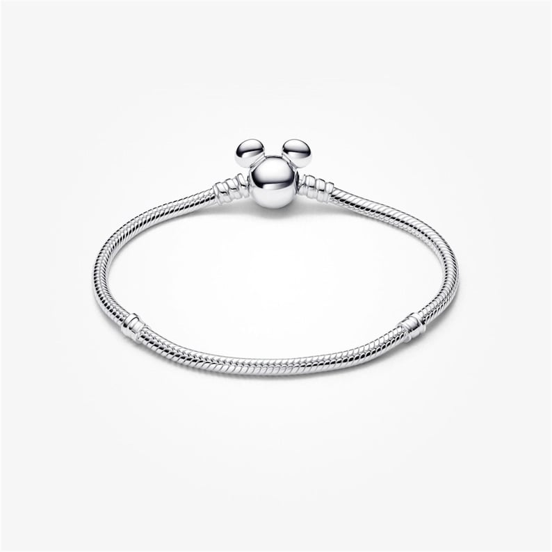 May include: A silver bracelet with a Mickey Mouse-shaped charm. The bracelet has a snake chain design and a round clasp. The charm features three round spheres, two for ears and one for the head. The bracelet is displayed on a white background.