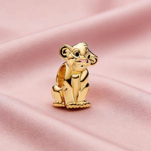 May include: A gold-coloured charm depicting a seated lion cub with a pink nose and large ears. The charm is highly detailed and has a polished finish. The charm is likely made of metal and is a collectible item.