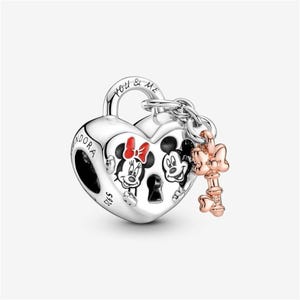 May include: A silver heart-shaped charm with Mickey and Minnie Mouse illustrations. The charm has a key and the words "You & Me". The key is rose gold. The charm is a Disney-themed jewelry item.