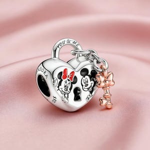 May include: A silver heart-shaped charm with Mickey and Minnie Mouse figures, a key, and the words "YOU & ME." The charm has a lock design with a key attached. Minnie Mouse has a red bow.