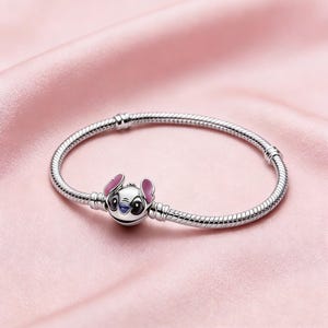 May include: A silver bracelet with a charm depicting Stitch from Lilo & Stitch. The charm has pink ears, blue eyes, and a white face. The bracelet is displayed on a pink satin background.