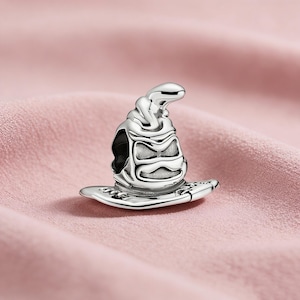 May include: A silver charm in the shape of a wizard's sorting hat. The hat is detailed with textured patterns and a wide brim. The charm is designed to be worn on a bracelet or necklace.