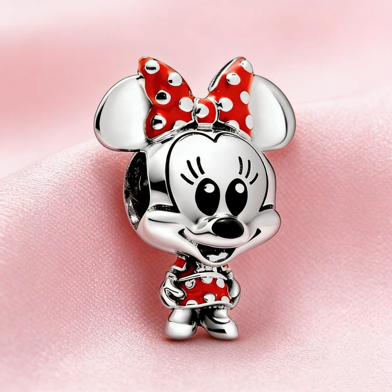 Puede incluir: Un charm plateado con un personaje de dibujos animados con un lazo y un vestido de lunares rojos. El charm tiene grandes ojos negros y una expresi&oacute;n sonriente. Las orejas del personaje son plateadas, sobre un fondo rosa.
