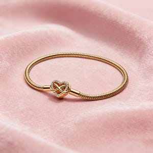 Könnte beinhalten: Ein goldfarbenes Armband mit herzförmigem Verschluss. Der Verschluss hat ein Unendlichkeitszeichen-Design, besetzt mit kleinen, klaren Edelsteinen. Das Armband liegt auf einem weichen, rosa Hintergrund.