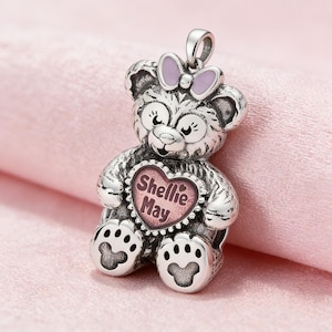 Könnte beinhalten: Silberfarbener Teddybär-Anhänger mit einem rosa herzförmigen Mittelteil mit der Aufschrift "Shellie May". Der Bär hat eine Schleife auf dem Kopf und Pfotenabdrücke auf den Füßen. Der Anhänger ist als Halskette konzipiert.